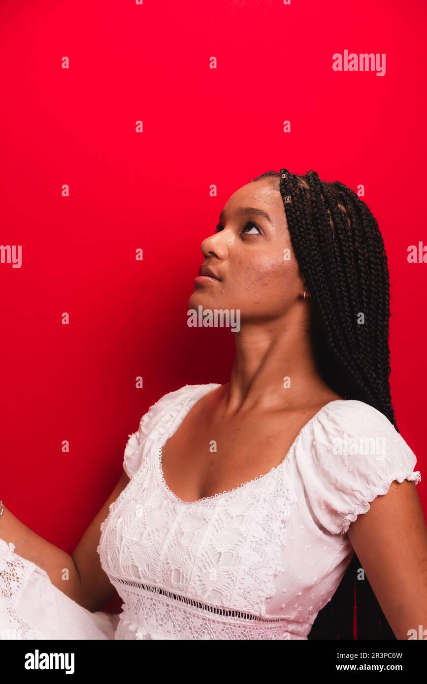 Schöne junge schwarze Frau, die sitzt und ruhig ist, weiße Kleidung trägt, mit langen Zöpfen im Haar. Isoliert auf rotem Hintergrund Stockfoto