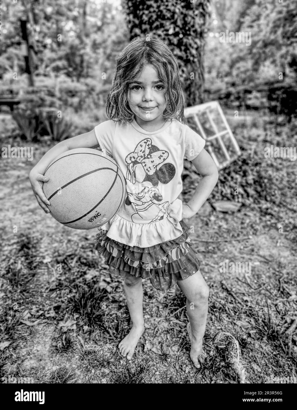 Ein kleines Mädchen, das draußen mit einem Basketball für die Kamera posiert. (Schwarzweißfoto) (NC-Status, 2023) Stockfoto