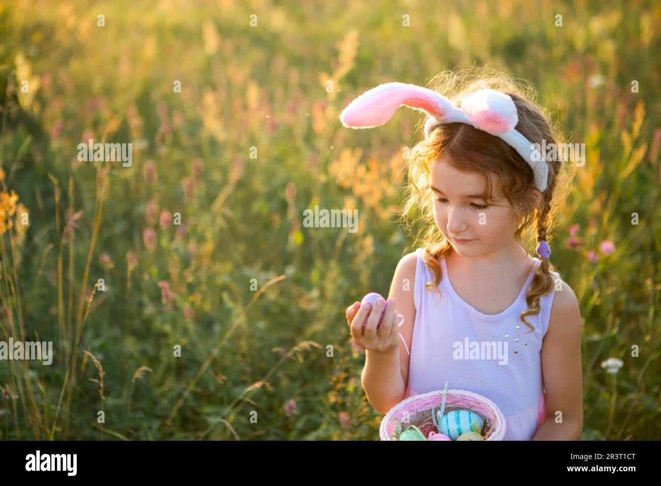 Süßes, lustiges Mädchen mit bemalten Ostereiern im Frühling in der Natur auf einem Feld mit goldenem Sonnenlicht und Blumen. Osterferien, Ostern Stockfoto