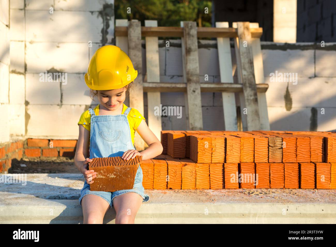 Ein kleines Mädchen mit gelbem Schutzhelm spielt Baumeister auf der Baustelle ihres zukünftigen Hauses. Erwartung, sich zu bewegen, zu wählen Stockfoto