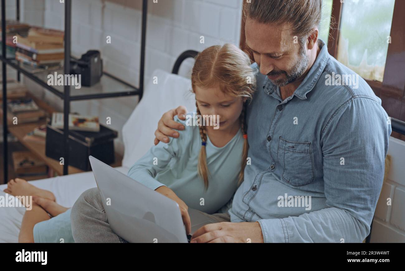 Vater benutzt Laptop-Computer mit der kleinen Tochter. Glückliche Familie, Vater entspannt sich und genießt es, zu Hause online zu arbeiten, während das kleine süße Mädchen im Bett liegt Stockfoto