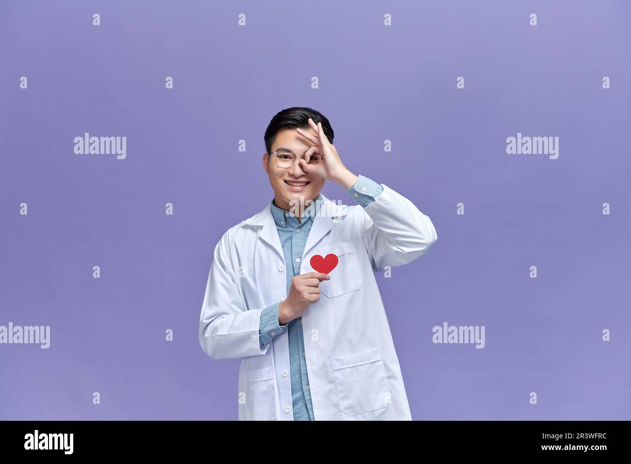 Ein Arzt, der ein Herz aus rotem Papier hält, mit einem fröhlichen Gesicht, das lächelt und das OK-Schild macht, mit einem Auge, das durch die Finger schaut Stockfoto