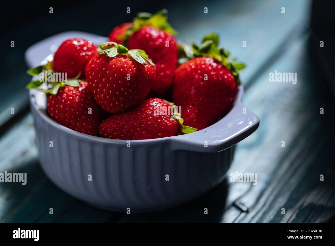 Eine einzelne Schüssel mit frischen Erdbeeren auf blauem Holzbrett in dramatischem Licht. Minimalszene Stockfoto