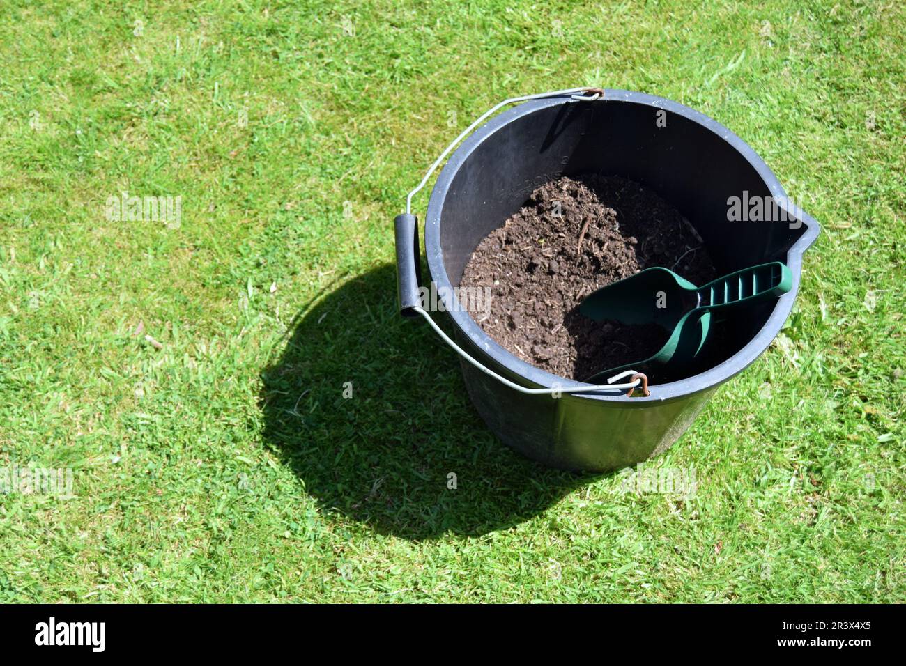 Eine grüne Kunststoffkelle befindet sich in einem schwarzen Kunststoffeimer gefüllt mit dunkelbraunem Kompost auf grünem Gras in einem Garten im Norden Englands. Stockfoto