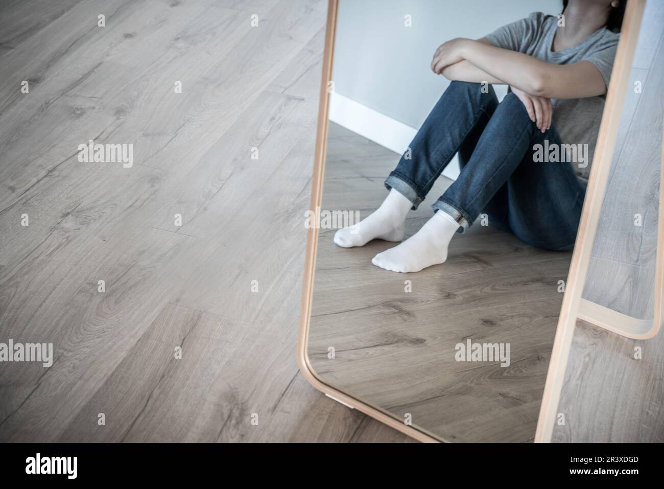 Reflexion im Spiegel einer traurigen Frau, die allein auf dem Boden sitzt, in einem leeren Raum, verzweifelt und einsam Konzept mit Kopierraum, Grauton Stockfoto