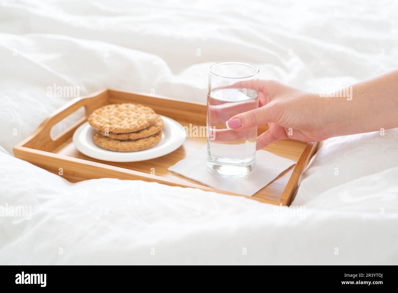 Weibliche Hand hält ein Glas Wasser über der Wanne mit Crackern auf dem Bett Stockfoto