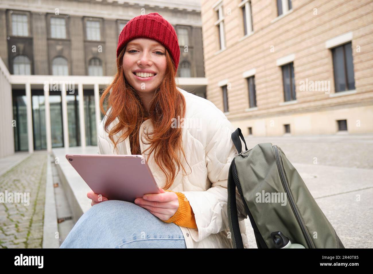 Das rothaarige Mädchen lächelt, sitzt im Freien in der Nähe des Gebäudes mit einem digitalen Tablet, Thermoskannen und Rucksack, stellt eine Verbindung zum öffentlichen Internet her und ist dabei auch gut Stockfoto