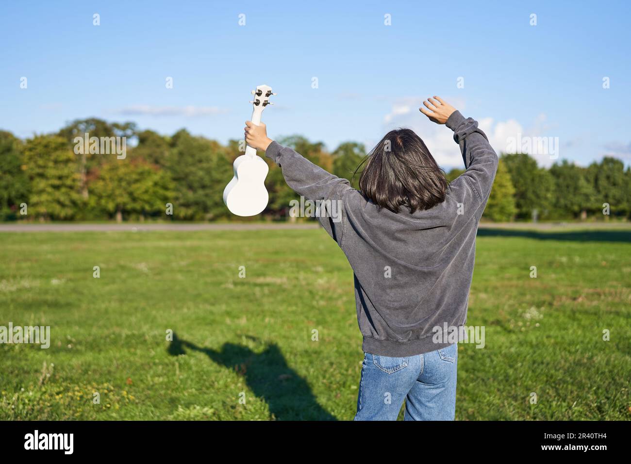 Fröhliche junge Frau, die mit ihrem Musikinstrument tanzt. Mädchen hebt ihre Ukulele hoch und posiert im Park auf grünem Feld Stockfoto