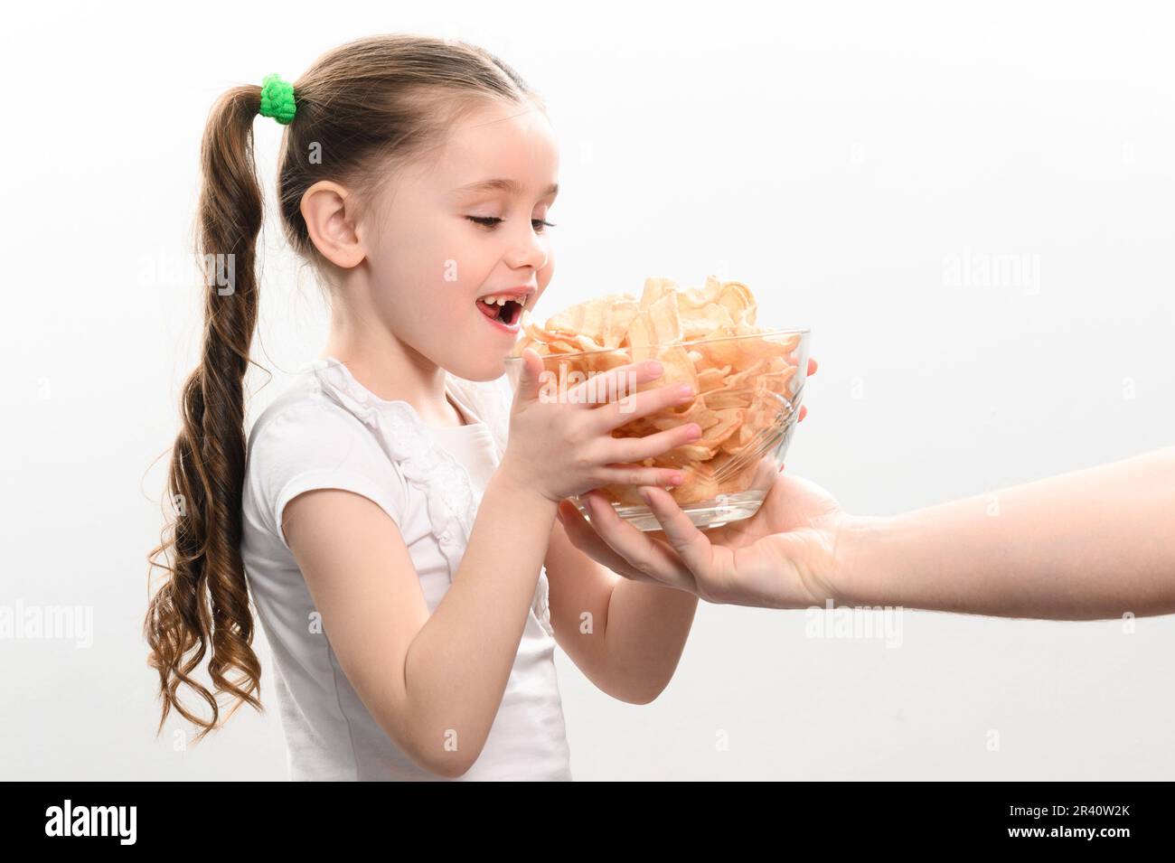 Das kleine Mädchen erhält eine große Schüssel Chips Snacks mit Schmalz, weißes Hintergrundporträt eines kleinen Mädchens, das Chips isst, ein Kind und ungesundes Fast Food. Stockfoto