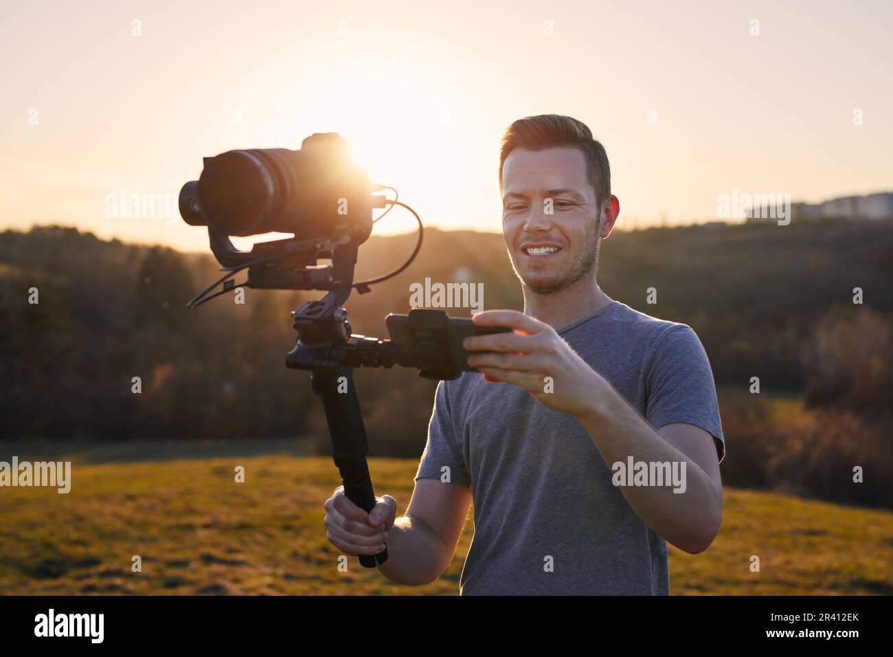 Lächelnder Mann beim Filmen mit Kamera und Gimbal. Porträt eines fröhlichen Videofilmers vor der Landschaft bei wunderschönem Sonnenuntergang. Stockfoto