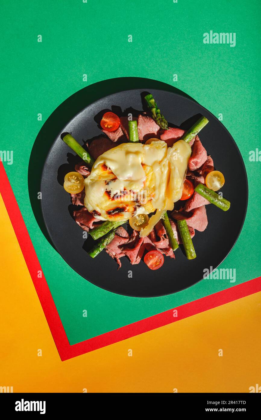 Köstlicher Raclette-Käse serviert über Rinderbraten mit Tomaten und Spargel, Gericht auf grünem Hintergrund. Hartes Licht, tiefer Schatten Stockfoto