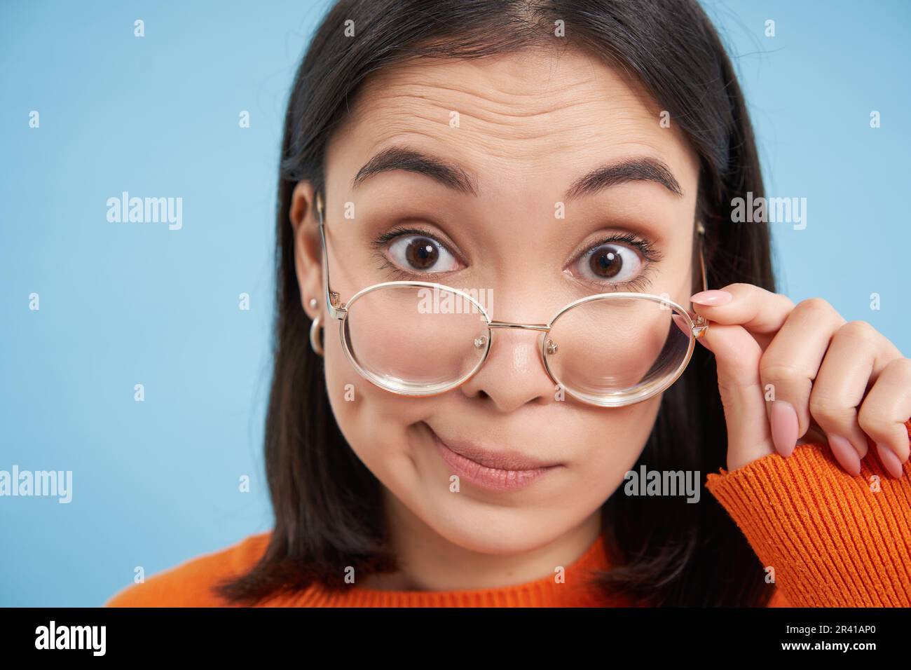 Nahaufnahme des überraschten Mädchens, schaut näher in die Kamera mit Brille, steht vor blauem Hintergrund Stockfoto