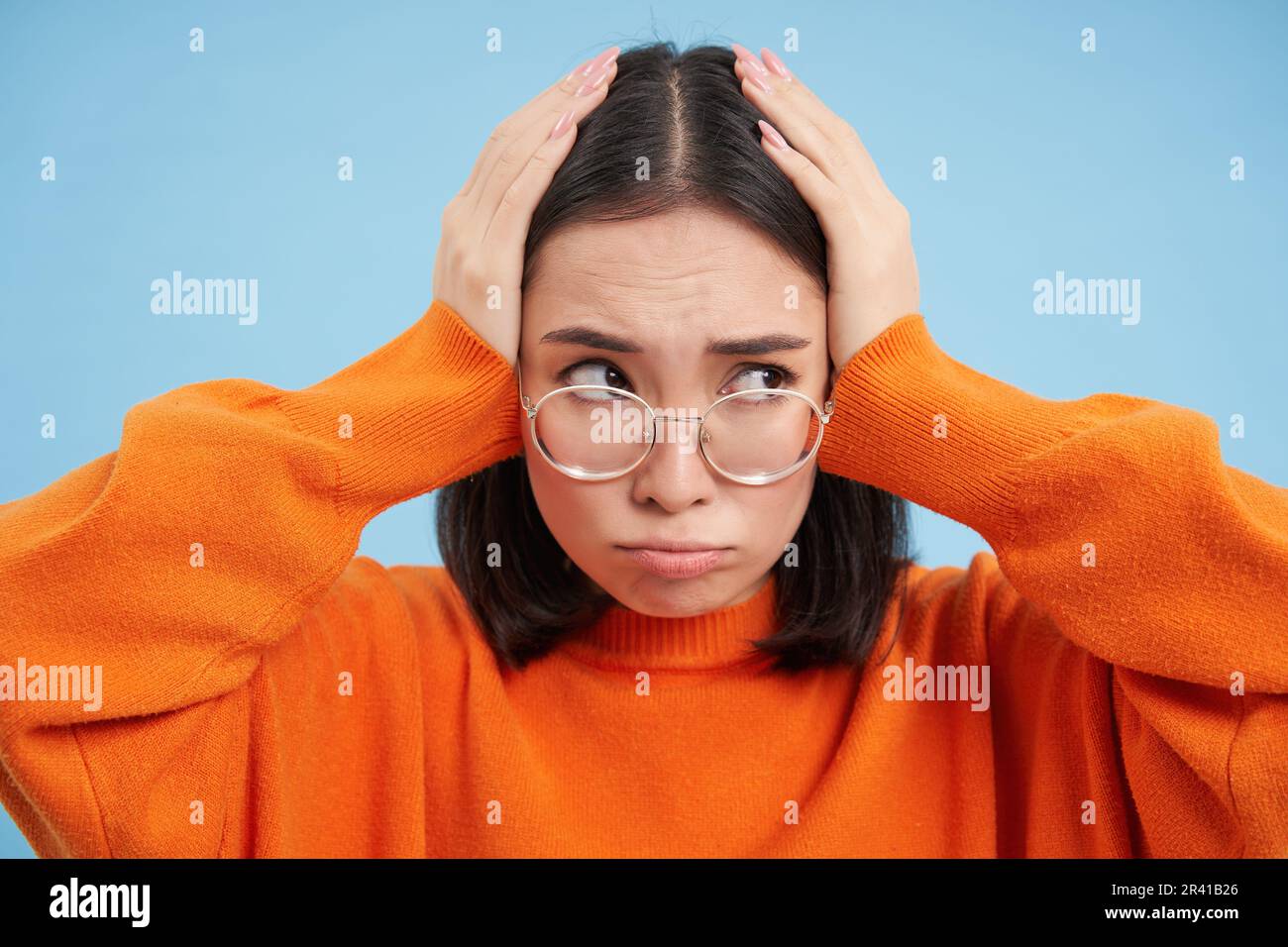 Frustrierte junge Frau mit Brille, hält Hände am Kopf, kompliziert, von Problemen beunruhigt, steht über blauem Hintergrund Stockfoto