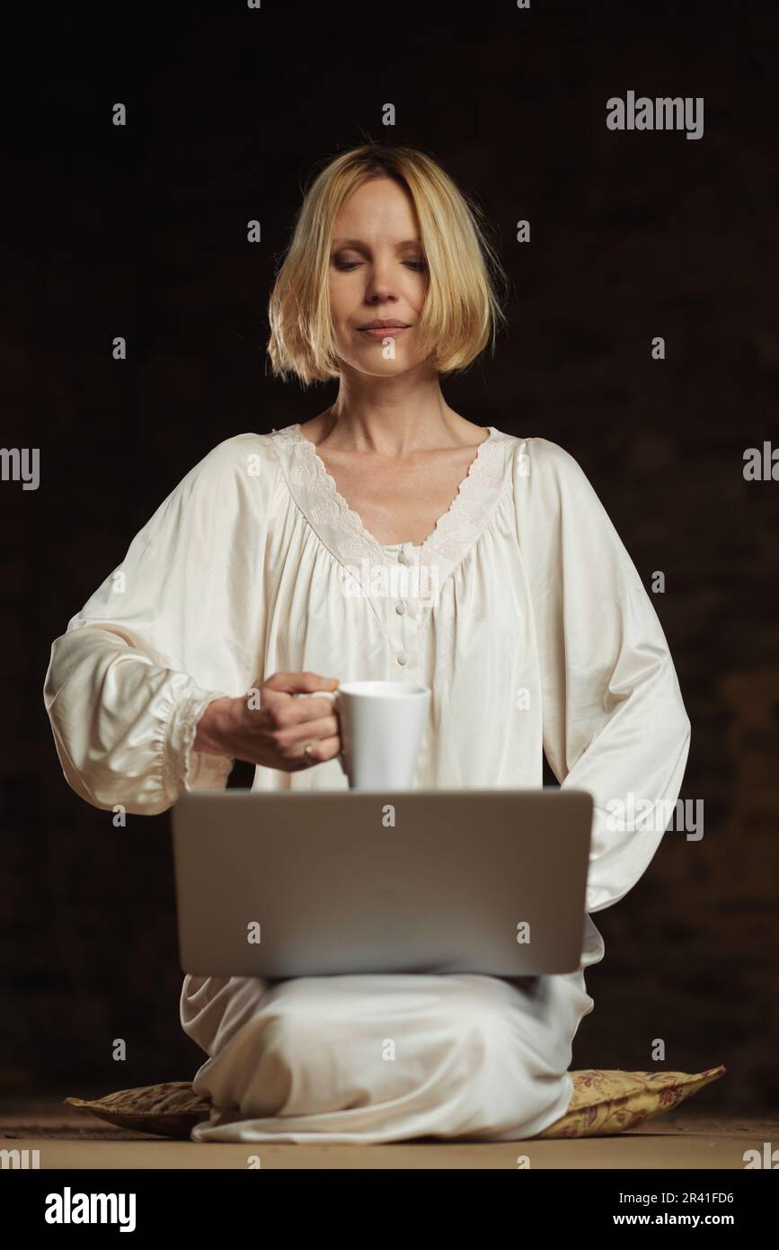 Frau mit Kaffee und Laptop. Workaholismus, Deadline-Konzept. Dunkler Hintergrund. Leeres Zimmer. Stockfoto