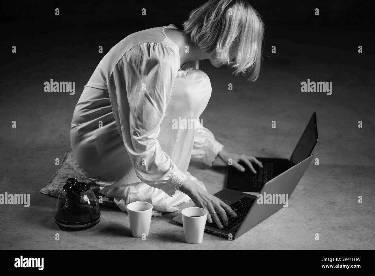 Frau mit Kaffeetasse vor dem Laptop. Leerer Raum, dunkler Hintergrund. Deadline, Recycling, Fleiß-Freelancer-Konzept. Schwarzweißbild. Stockfoto