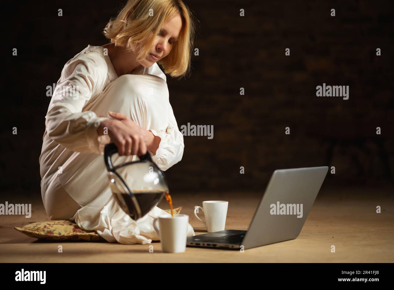 Eine Frau gießt Kaffee ein, während sie vor einem Laptop sitzt. Leerer Raum, dunkler Hintergrund. Deadline, Recycling, Fleiß-Freelancer-Konzept. Stockfoto