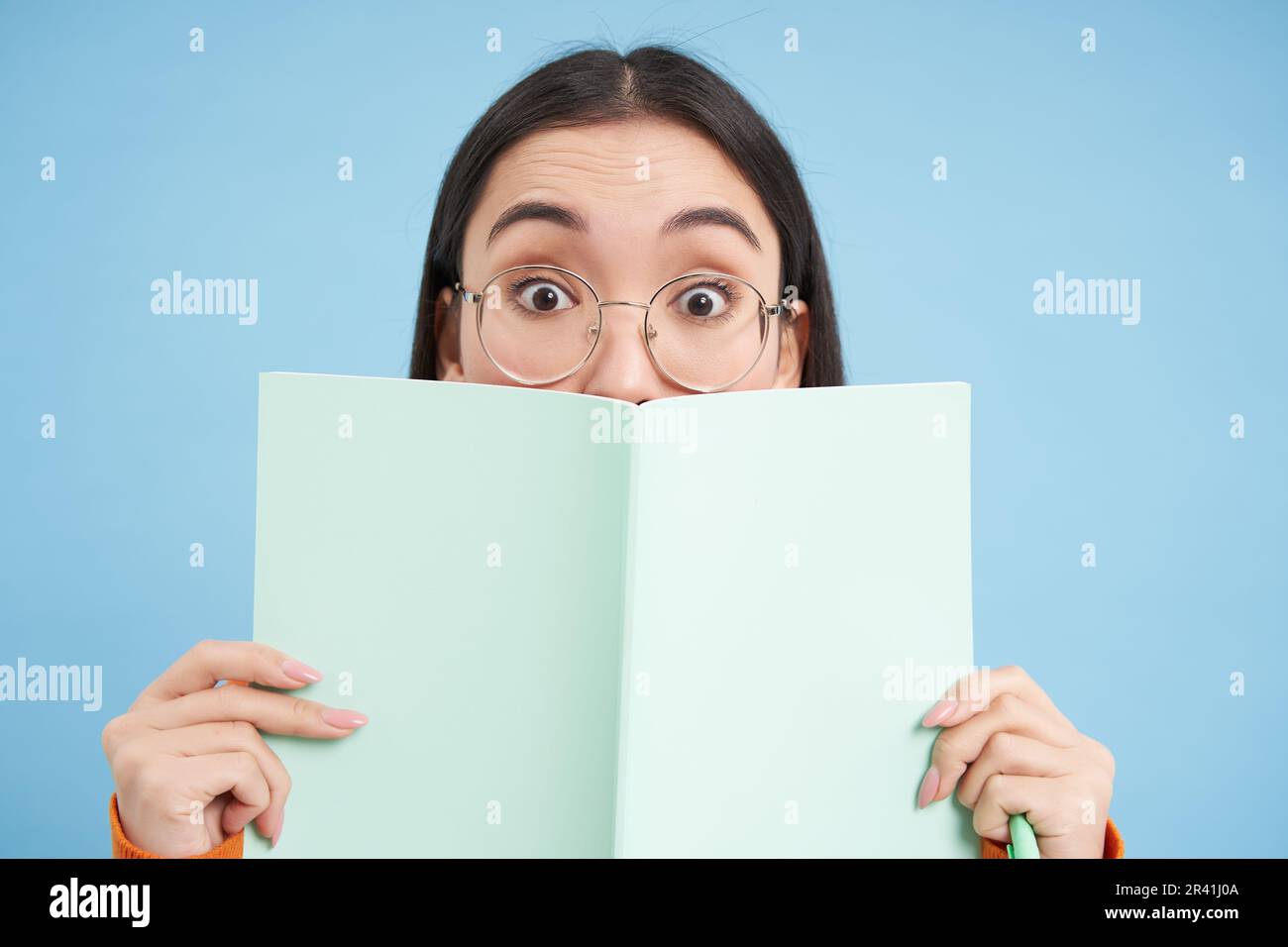 Süße asiatische Frau in Brille, versteckt ihr Gesicht hinter dem Notizbuch und lächelt mit Augen, neugieriger Blick, steht auf blauem Hintergrund. Stu Stockfoto