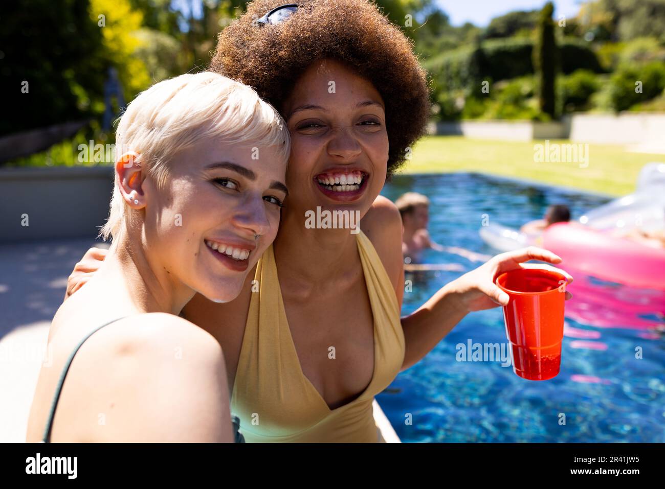 Porträt von fröhlichen, vielfältigen Freundinnen, die eine Poolparty veranstalten, sich umarmen und im Garten lächeln Stockfoto