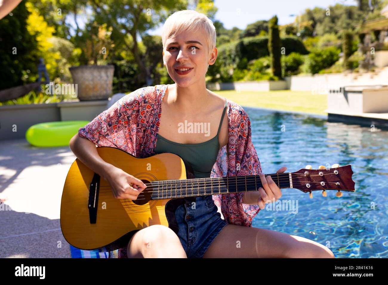 Porträt einer glücklichen Kaukasierin, die eine Poolparty veranstaltet, Gitarre spielt und im Garten lächelt Stockfoto