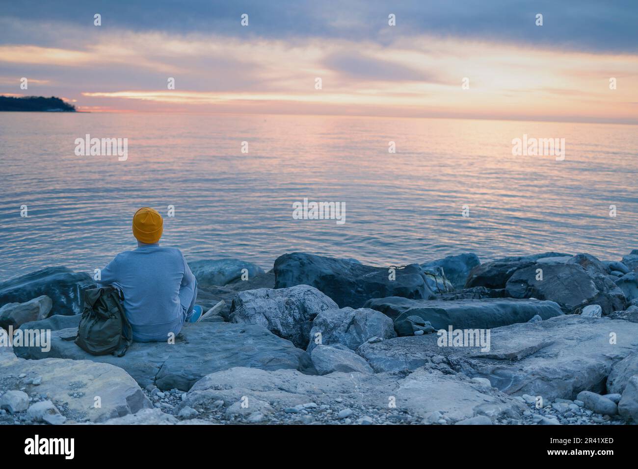 Ein Aukasier mit blauem Pullover und Hut hält seinen Rucksack und schaut auf das ruhige Meer. Stockfoto