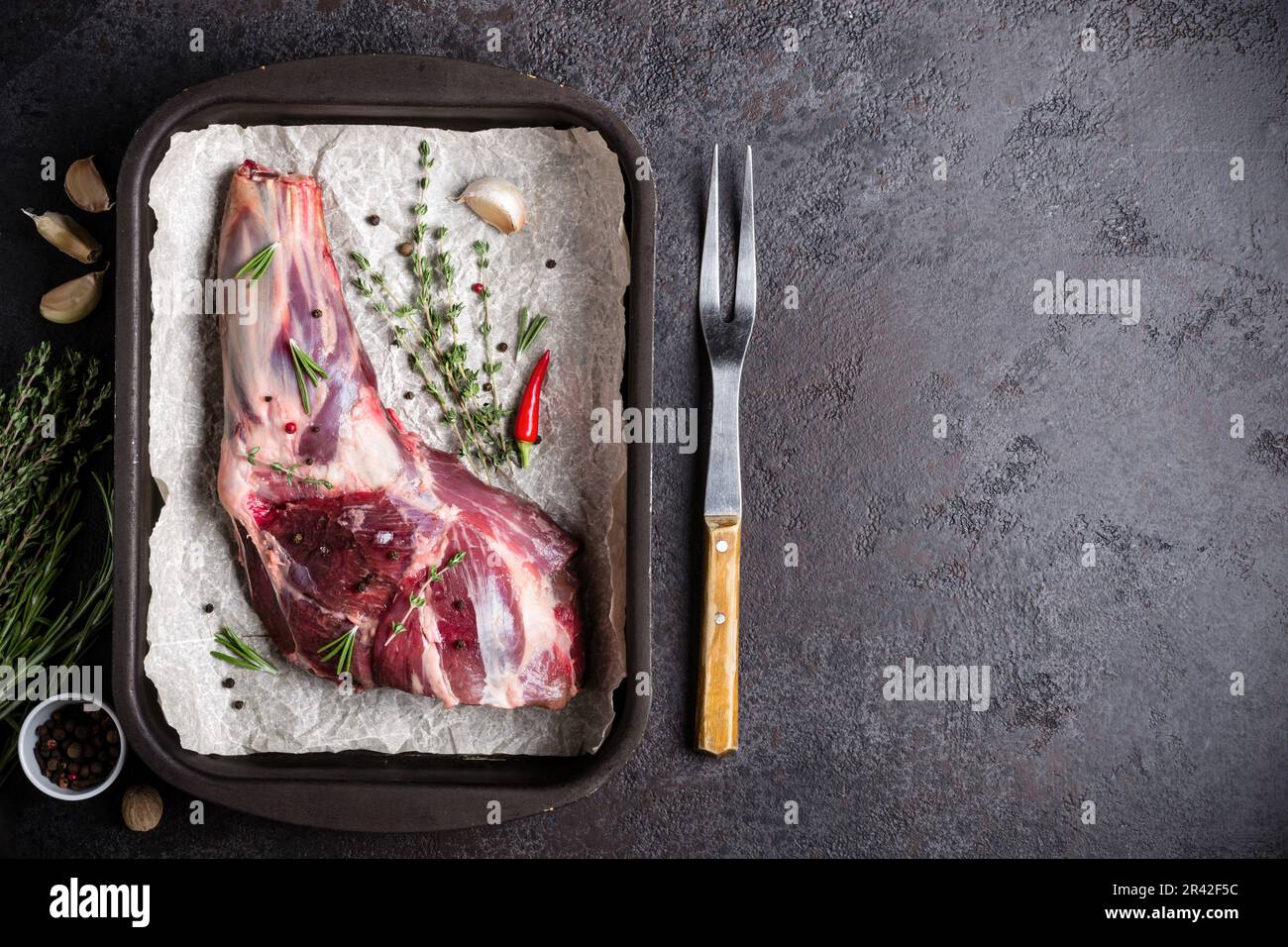 Rohes, frisches Lammfleisch, Kräuter und Gabel auf schwarzem Steinhintergrund Stockfoto