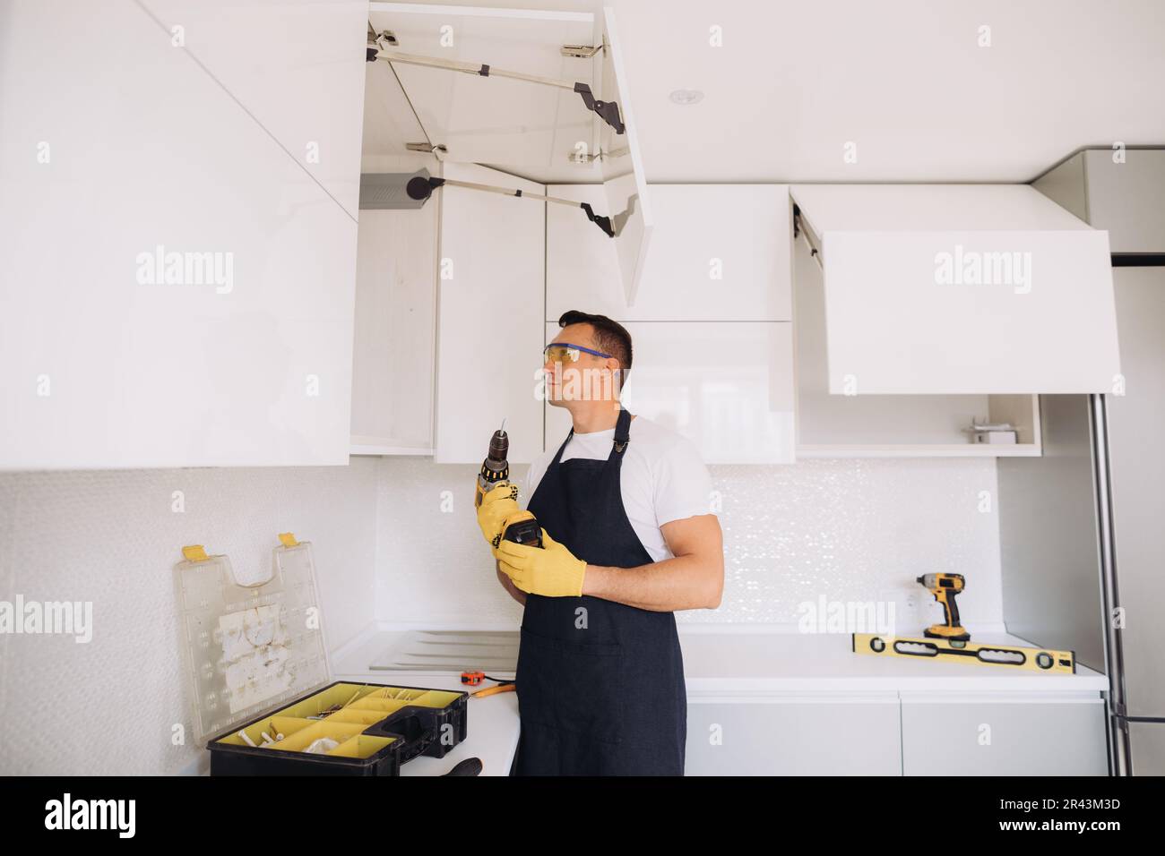 Ein Hausmeister, der Küchenmöbel in einer schwarzen Schürze und gelben Handschuhen mit einem Power-Skrewdriver installiert Stockfoto