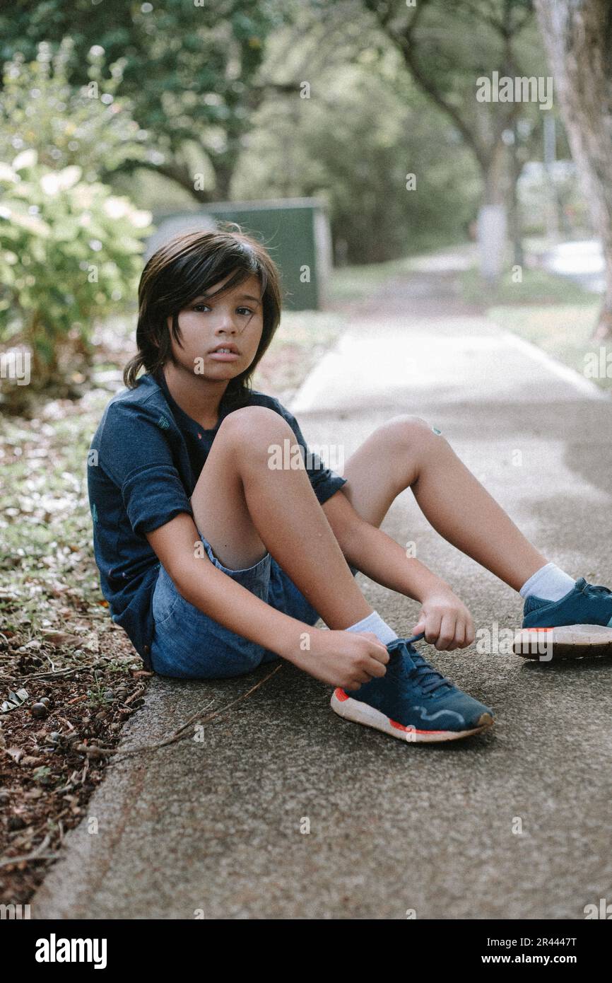 Ein junger, multiethnischer asiatischer Junge, der auf dem Bürgersteig sitzt und seine Schuhe bindet. Stockfoto