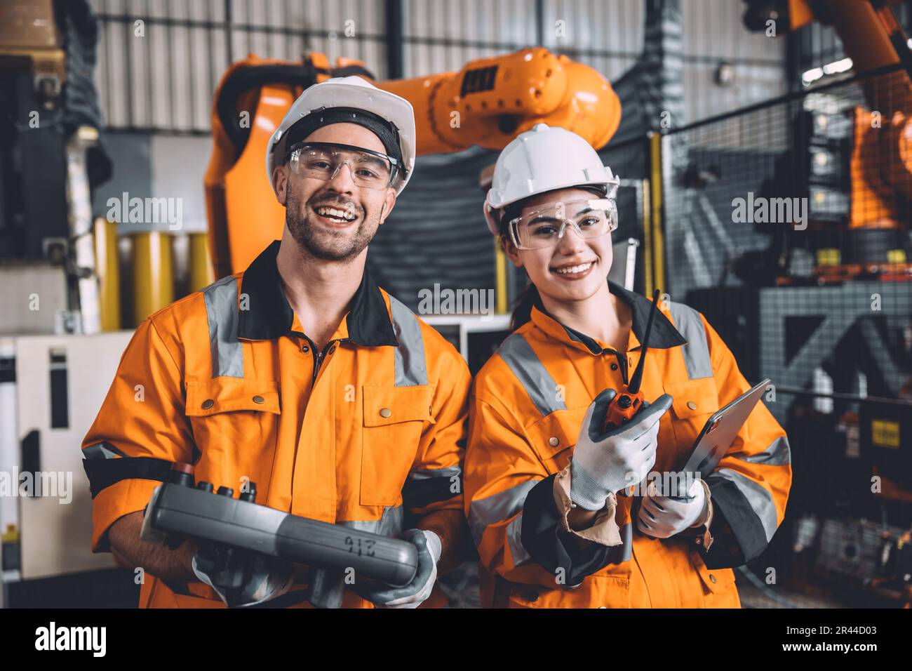 Glücklicher junger Ingenieur, der in der Automatisierungsfabrik mit dem Serviceteam beim Roboterschweißen zusammenarbeitet. Lächelnder Mann und lächelnde Arbeiterin im Sicherheitsanzug Arbeiter Stockfoto