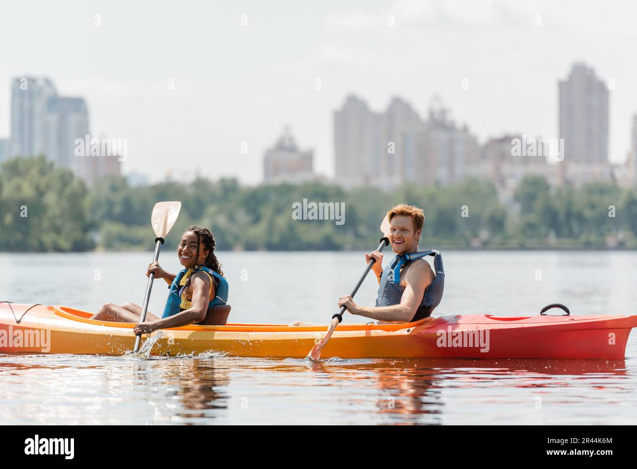 Eine bezaubernde afroamerikanische Frau und ein sportlicher rothaariger Mann in Schwimmwesten, der beim Segeln im Kajak am Flussufer mit verschwommener Stadt auf die Kamera schaut Stockfoto