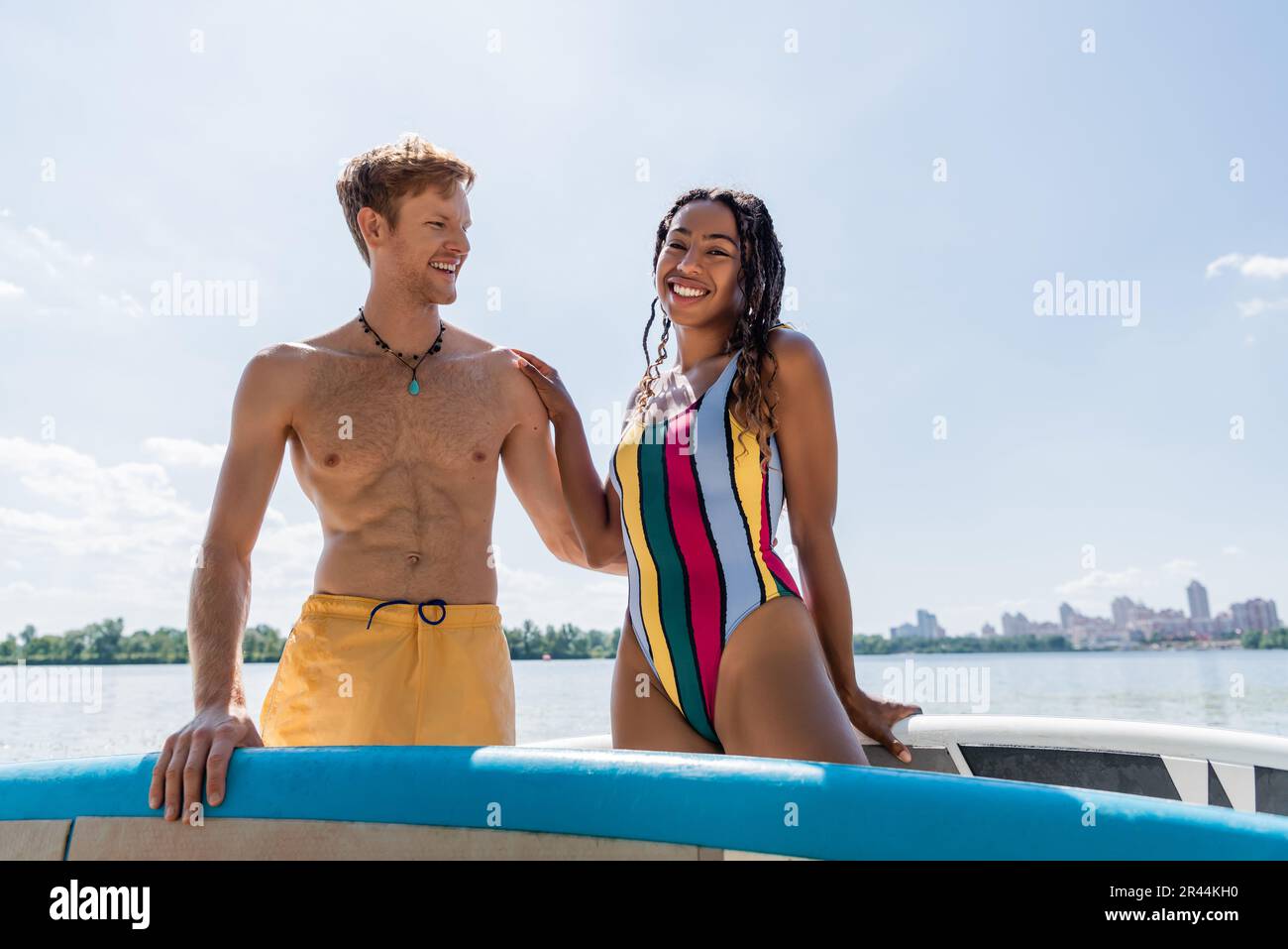 Eine fröhliche und bezaubernde afroamerikanische Frau in bunten Badeanzügen, die in der Nähe eines jungen rothaarigen Mannes in die Kamera schaut, und draußen unter blauem Himmel ein Sup-Board Stockfoto