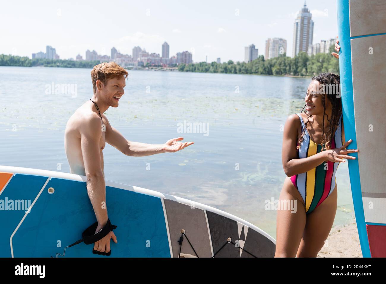 Glücklicher rothaariger Mann mit Sup-Board und Gespräch mit einer bezaubernden afroamerikanischen Frau in farbenfrohen Badeanzügen in der Nähe des Sees und der Stadtlandschaft im Hintergrund Stockfoto