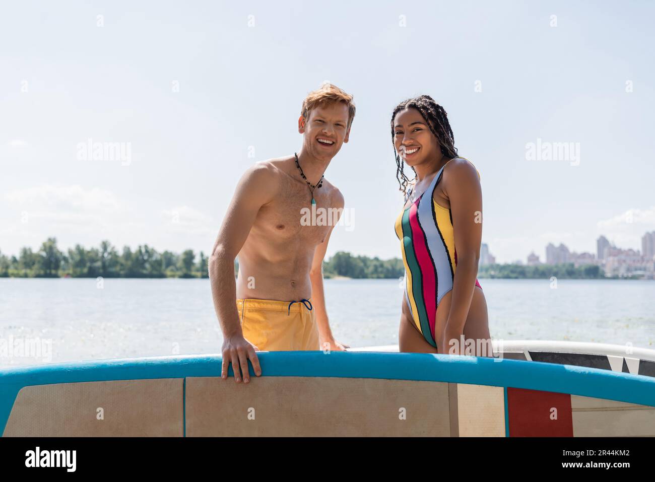 Eine afroamerikanische Frau im gestreiften Badeanzug und ein junger rothaariger Mann in gelben Badehosen, der in der Nähe von Sup Boards mit See und Stadt auf die Kamera schaut Stockfoto