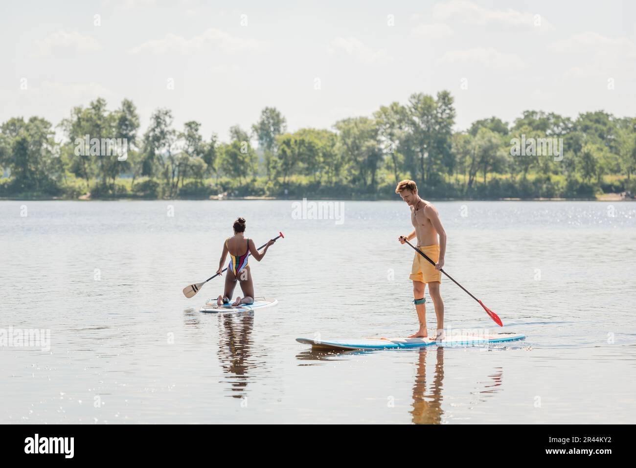 Junger und sportlicher rothaariger Mann, der auf dem Sup-Board steht, mit einem Paddel in der Nähe einer afroamerikanischen Frau in bunten Badeanzügen, die auf dem malerischen See mit Gre segelt Stockfoto