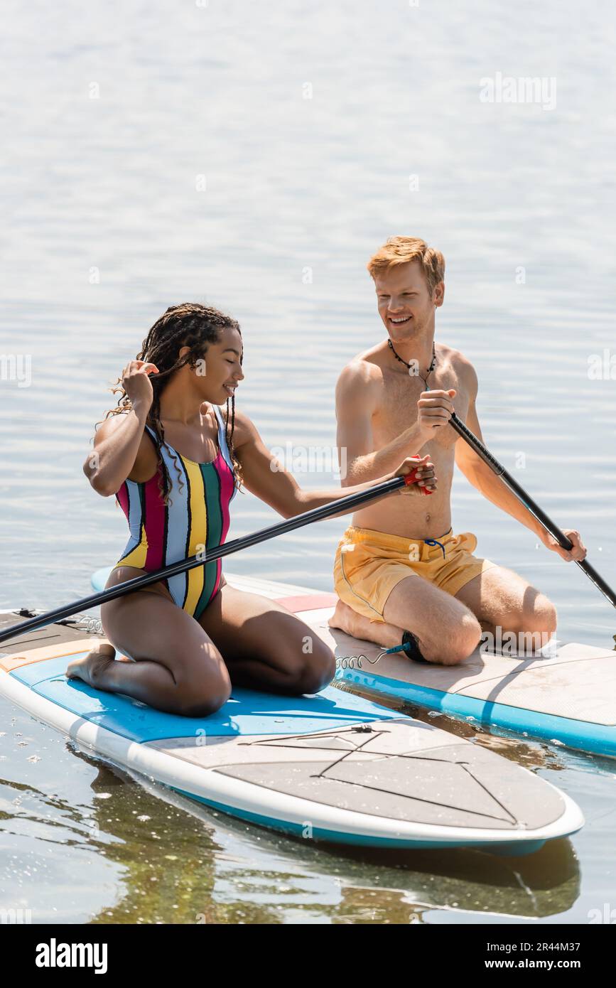 Ein glücklicher rothaariger Mann, der eine bezaubernde afroamerikanische Frau in bunten Badeanzügen ansieht, während er auf dem Sup-Board segelt und gleichzeitig Zeit zusammen auf dem See verbringt Stockfoto