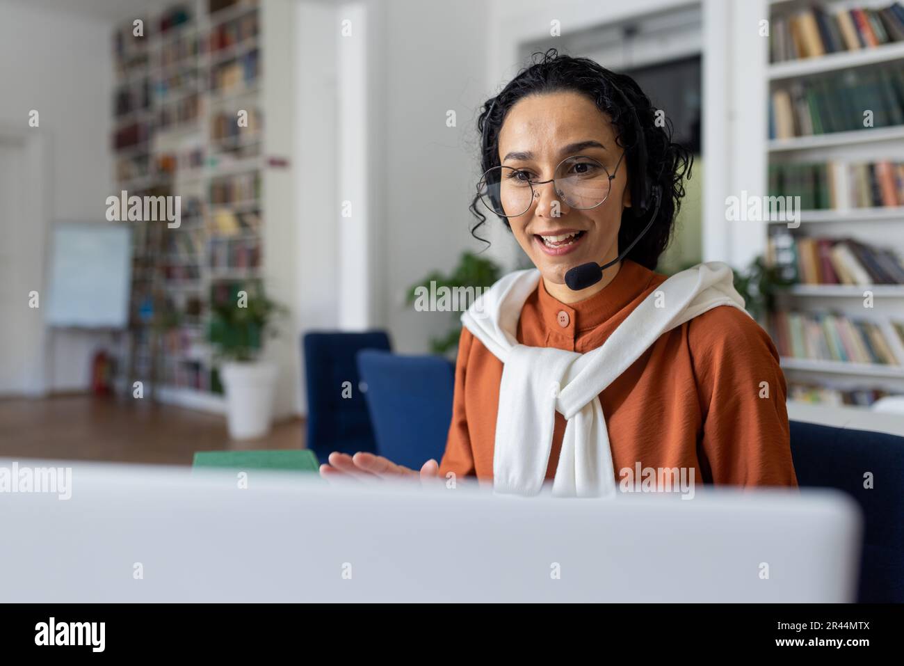 Lehrer führt Unterricht online durch, Frau mit Headset für Videogespräche lächelt und erklärt Vorlesungen online mit Laptop für Fernkommunikation und Lernen, sitzt am Schreibtisch in der Universitätsbibliothek am Schreibtisch. Stockfoto