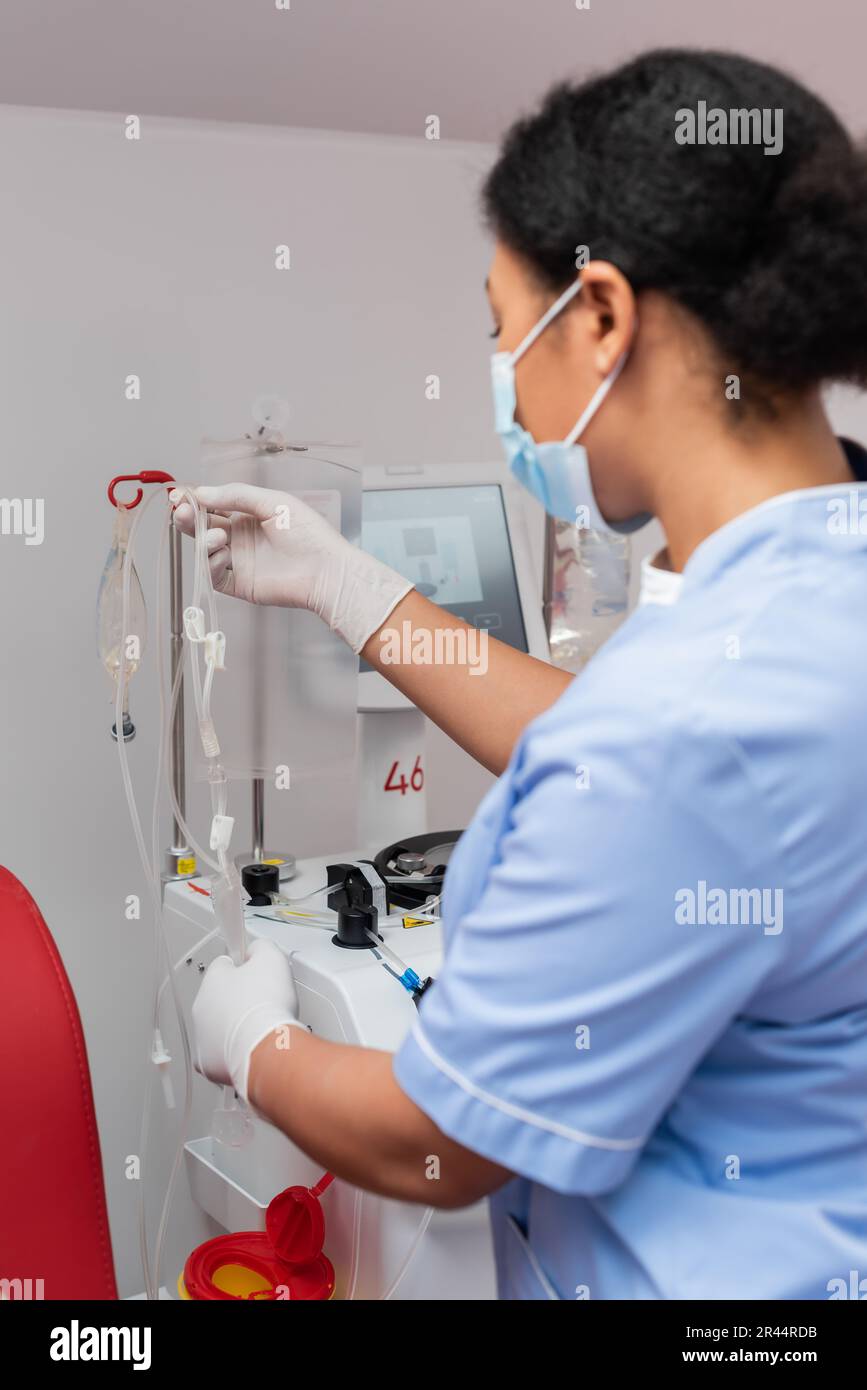 Multiethnische Krankenschwester in blauer Uniform und Latexhandschuhe, die das Bluttransfusions-Set in der Nähe eines Tropfstativs mit Infusionsbeuteln im Labor halten, verschwommener Vordergrund Stockfoto
