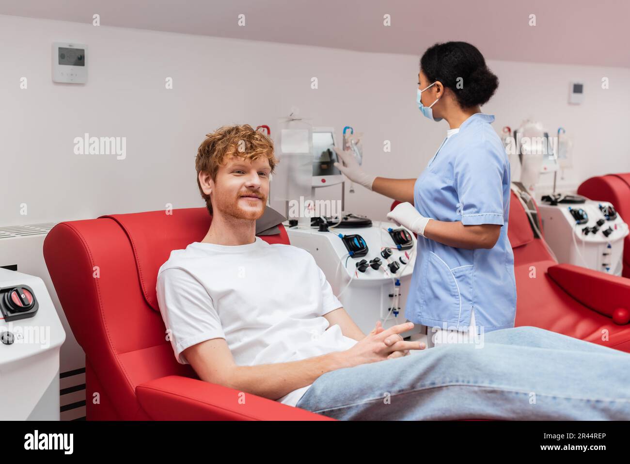 Multirassische Krankenschwester in medizinischer Maske und Latexhandschuhe, die Transfusionsgerät bedienen, nahe zufriedener rothaariger Mann, der auf einem bequemen Stuhl im Blut donati sitzt Stockfoto
