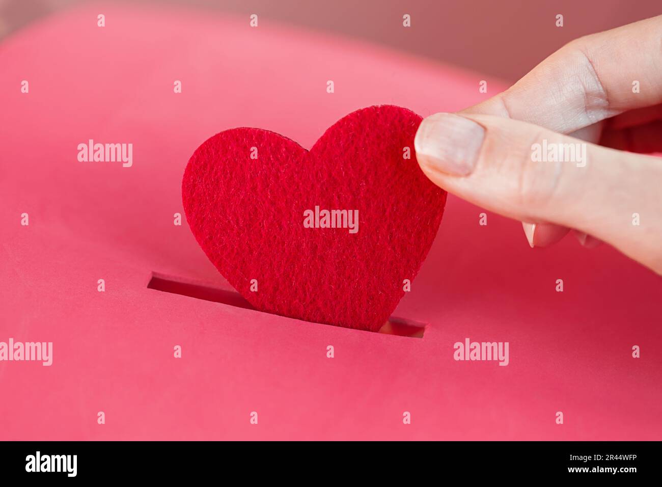 Die weibliche Hand steckt Red Heart in die rote Spendenbox. Konzept der aufrichtigen Hingabe zum Glauben. Konzept der Spende, Lebensrettung oder Wohltätigkeit. Nahaufnahme Stockfoto