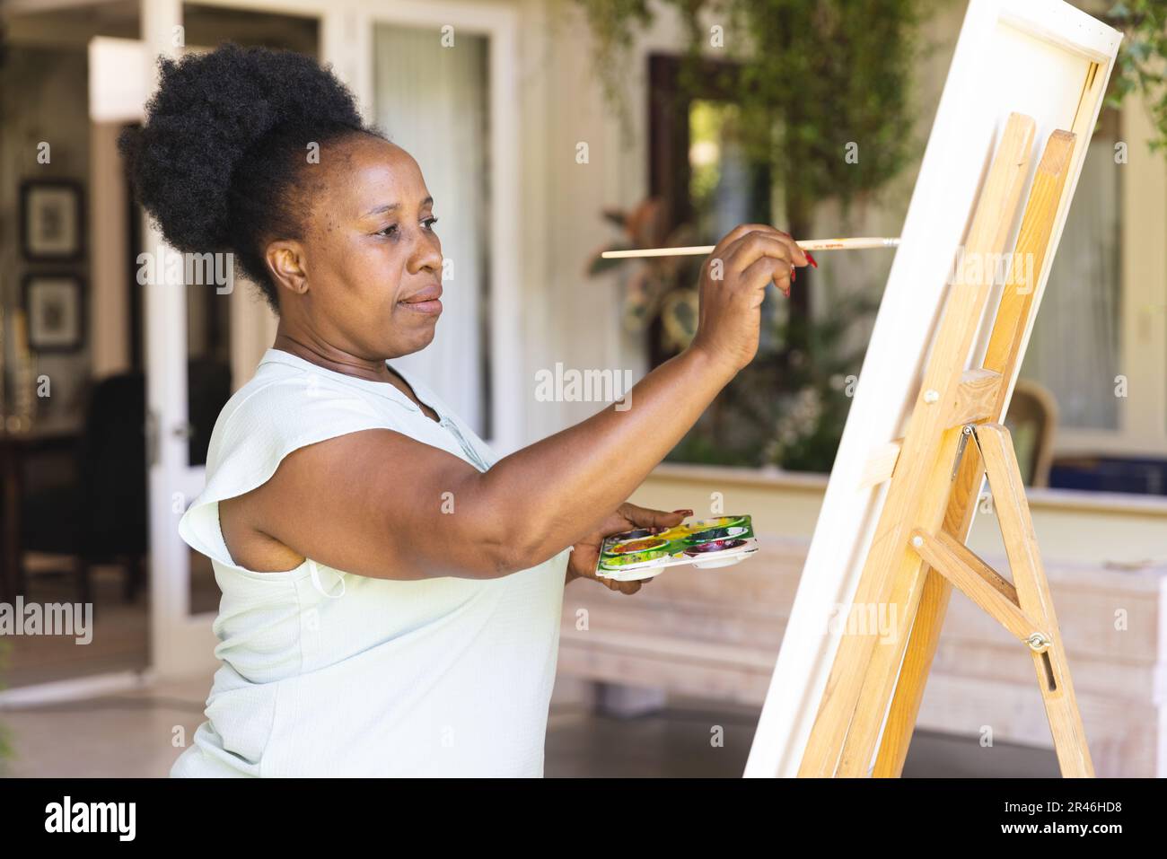 Fokussierte, glückliche, ältere afroamerikanische Frau, die zu Hause Leinwand mit Farbpaletten auf Staffelei hält Stockfoto