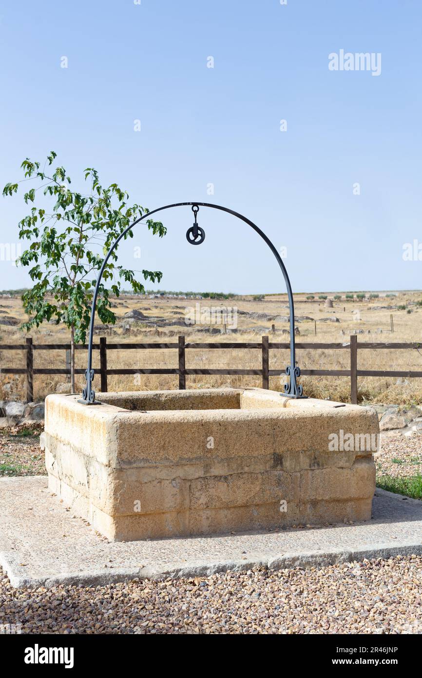 Steinbrunnen für Wasser in der Mitte des Feldes. Mit Metallbogen. Es ist auf einer Zementplattform. Hinter ihm ist ein kleiner Baum. Der Himmel ist blau. Vertikal. Stockfoto