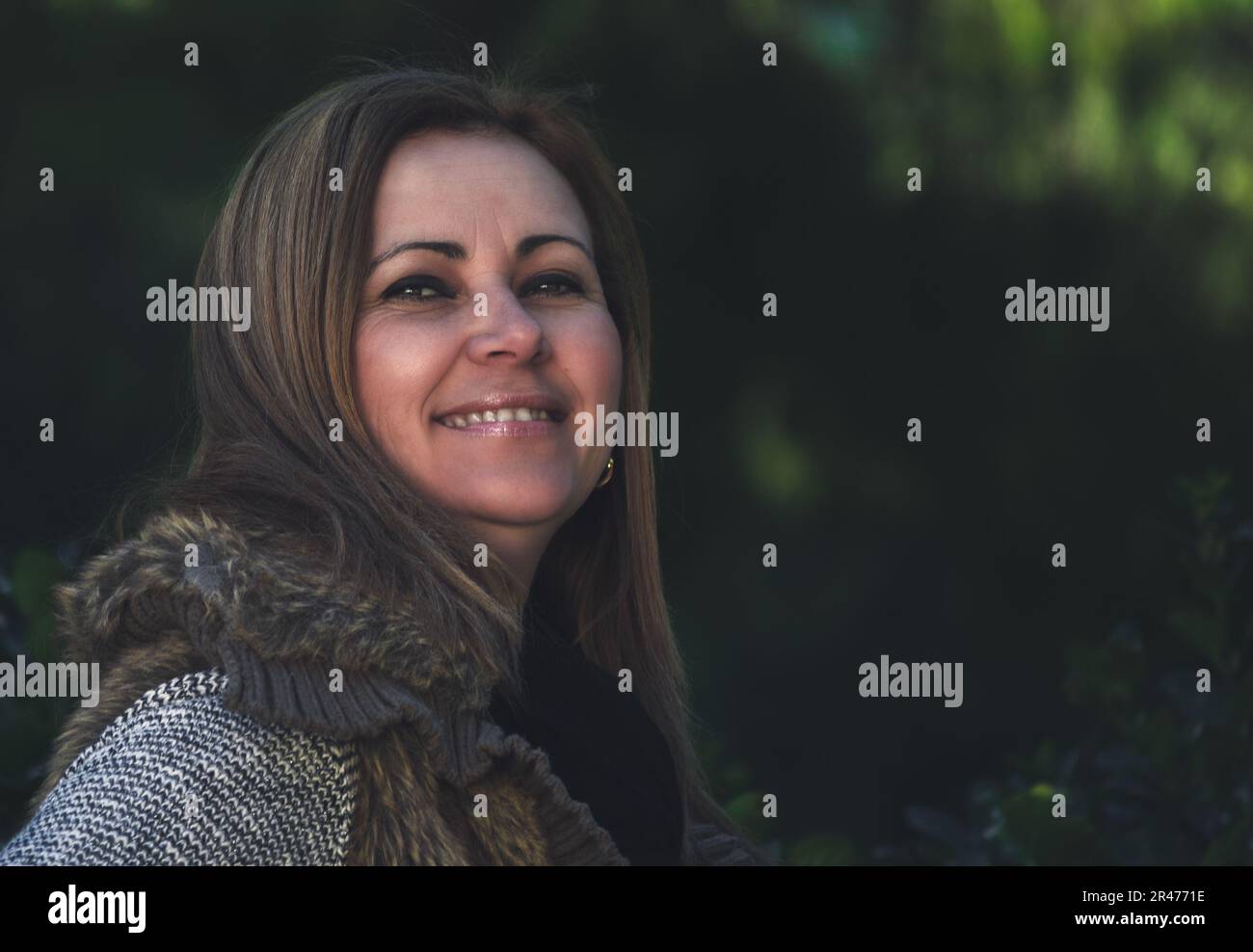 Porträt einer lächelnden blonden Frau mittleren Alters in der Natur, mit unscharfem dunkelgrünen Hintergrund. Lifestyle-Konzept. Stockfoto