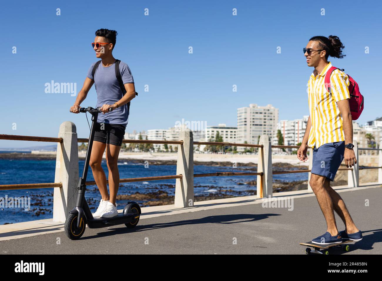 Fröhliches, birassisches, schwules Paar, das auf dem Roller und Skateboard auf der Promenade am Meer sitzt Stockfoto