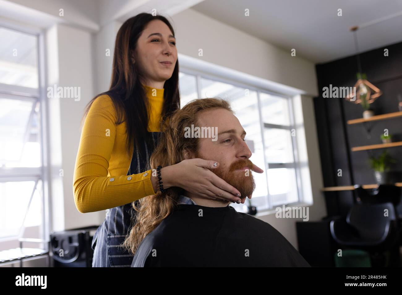 Fröhliche, weiße Friseurin mit Bart eines langhaarigen männlichen Klienten im Friseursalon Stockfoto