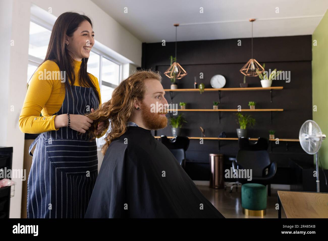 Glückliche weiße Friseurin, die Haare eines langhaarigen männlichen Klienten im Friseursalon hält Stockfoto