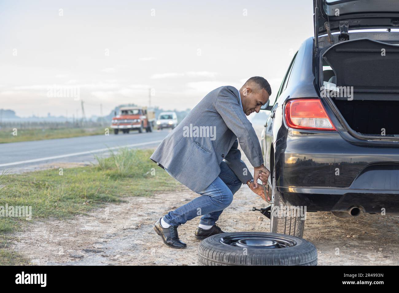 Ein Latino-Mann im Anzug wechselt einen Platten Reifen an seinem Auto am Straßenrand mit Kopierraum. Stockfoto