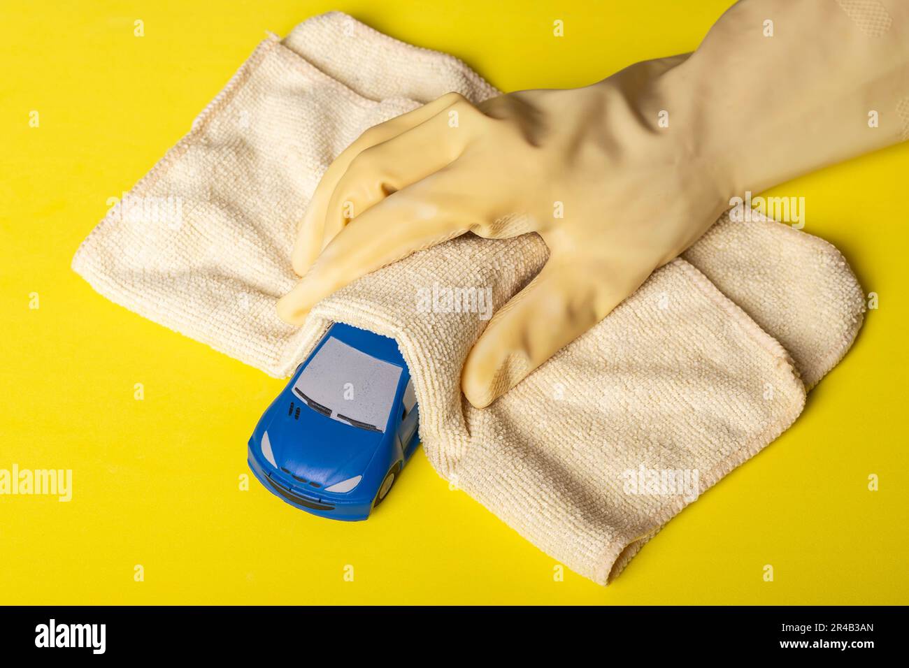 Ein blaues Spielzeugauto wird mit einer Hand gereinigt und desinfiziert, die mit einem gelben Handschuh und einem Mikrofasertuch auf gelbem Hintergrund bedeckt ist Stockfoto