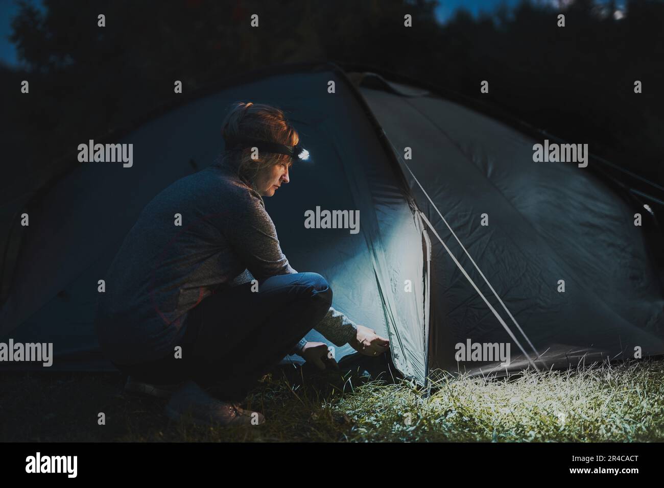 Sommerferien. Frau, die das Zelt am Abend beim Camping mit einer Stirnlampe eröffnet. Sommerausflug. Campingplatz vorbereiten. Urlaub im Freien Stockfoto