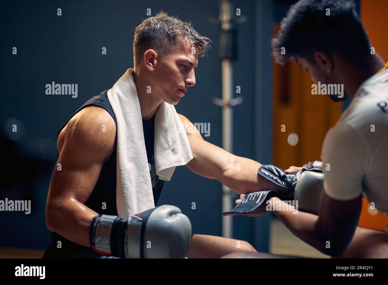 Junger muskulöser Boxspieler, der sich nach dem Spiel in der Garderobe ausruht, wobei der Trainer seine Handschuhe auszieht. Sport, Kampfsport, Lifestyle-Konzept. Stockfoto