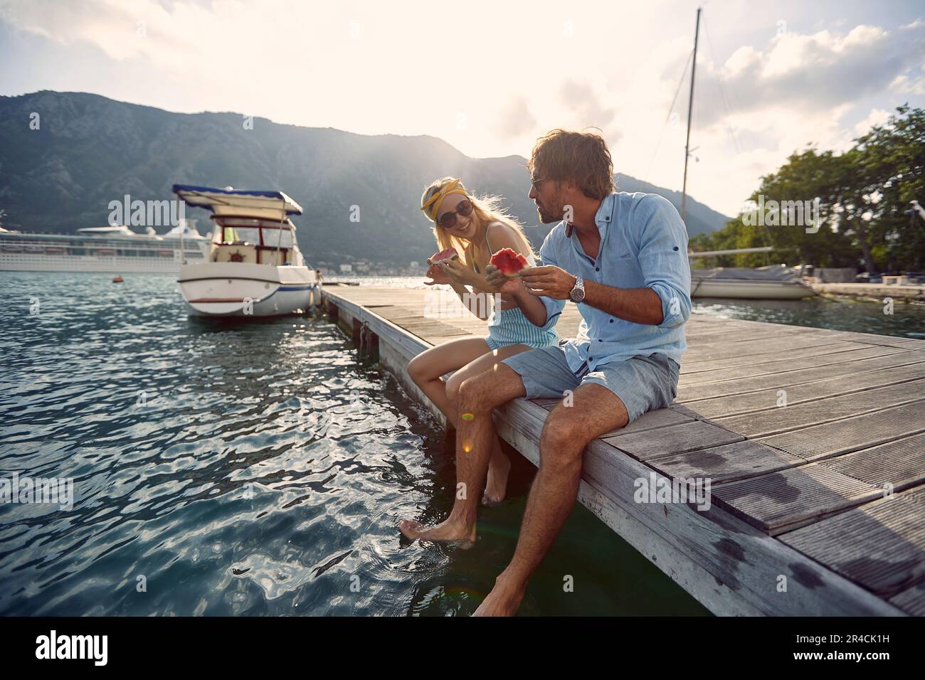 Mann und Frau sitzen auf dem Seepier, baumeln Beine und essen Wassermelone zusammen Stockfoto