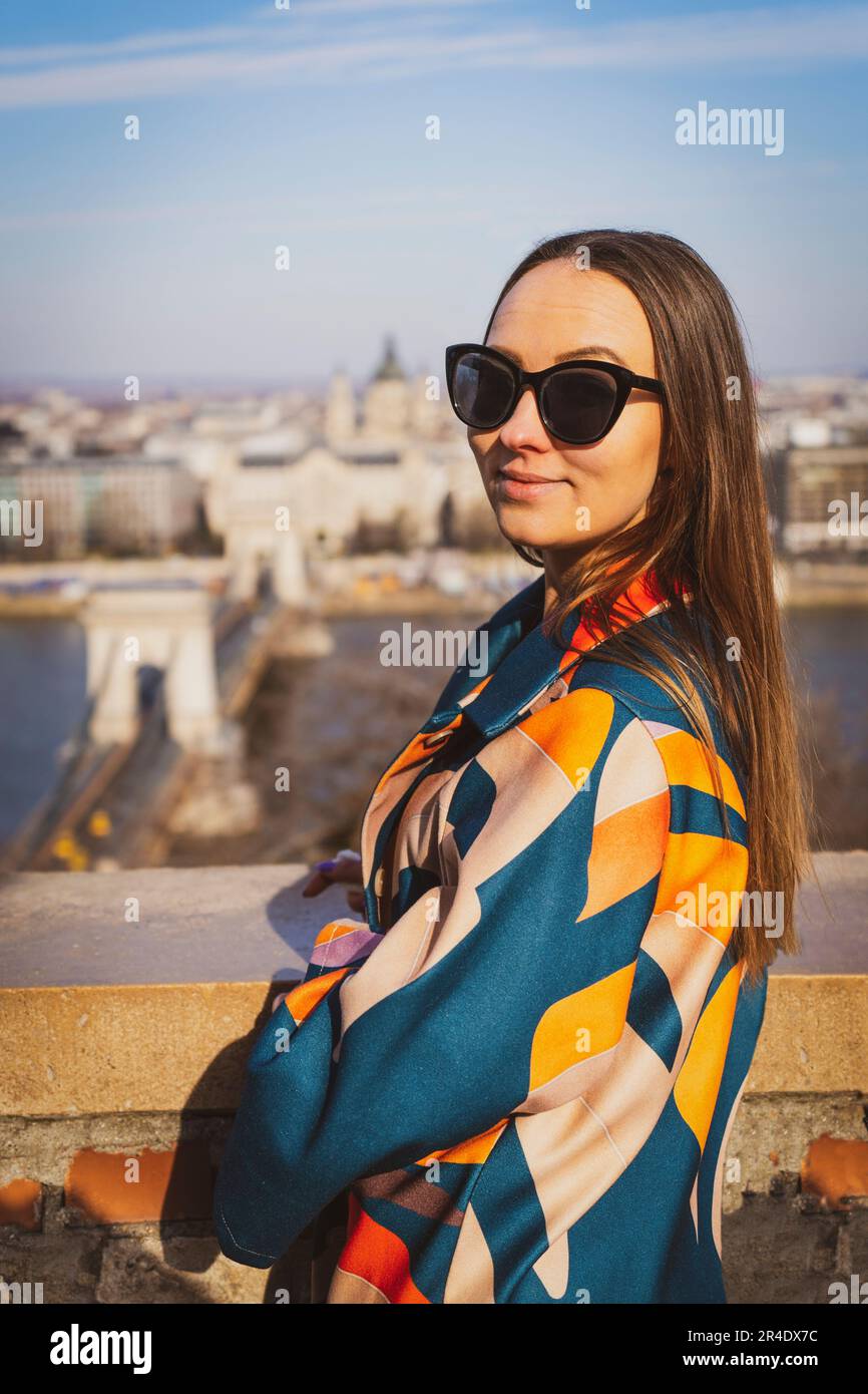 Junge Frau mit Sonnenbrille in einer farbenfrohen Jacke mit Budapest im Hintergrund Stockfoto